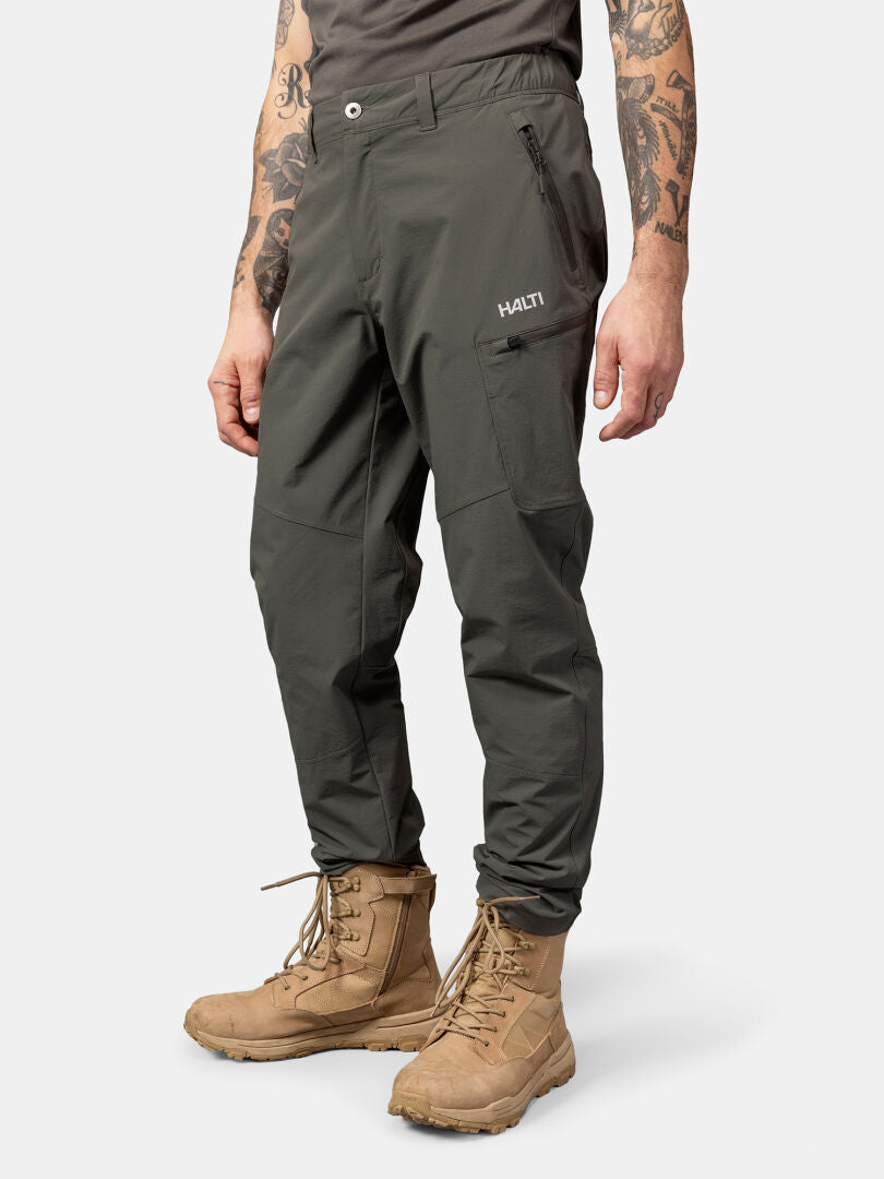 Person wears Halti Pallas Evo X-stretch Pants in olive green with zippered pockets and tan boots against a plain background.
