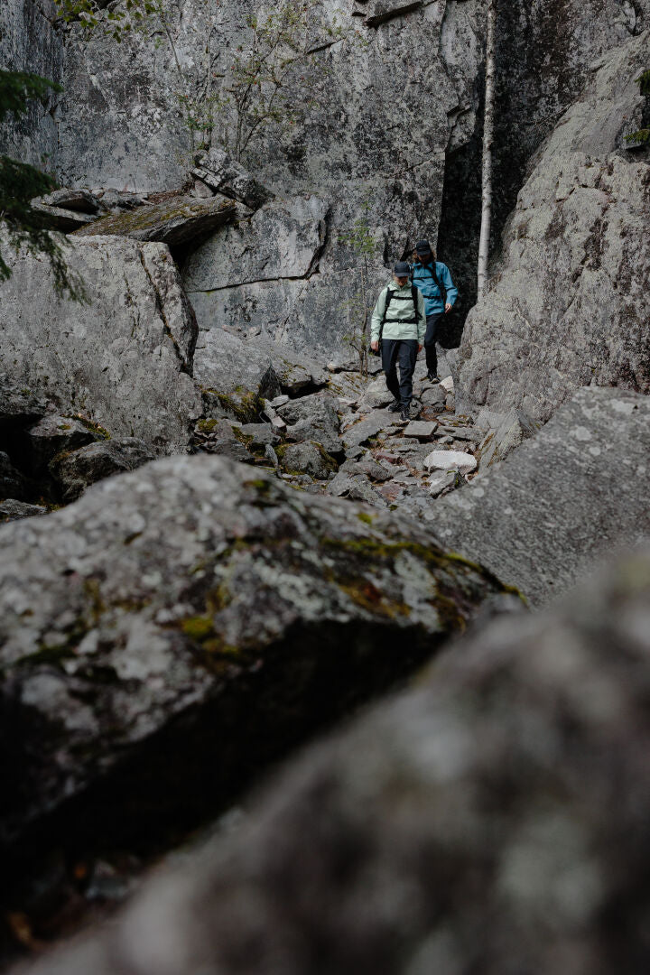 Two people in Halti Pisarat 2,5L DX Shell Pants hike a rocky forest trail beside a gray cliff.