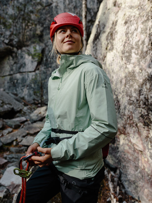 A person in a Halti Pisarat 2.5L DX Shell Jacket Womens and red helmet stands by a rocky wall with climbing gear.