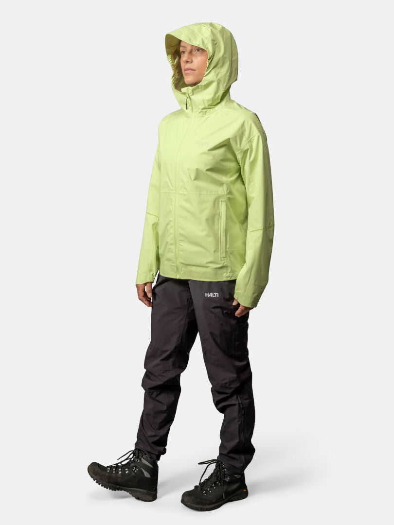 Woman wearing a Halti Pisarat 2,5L DX Shell Jacket in light green, black pants, and hiking boots, against a plain background.