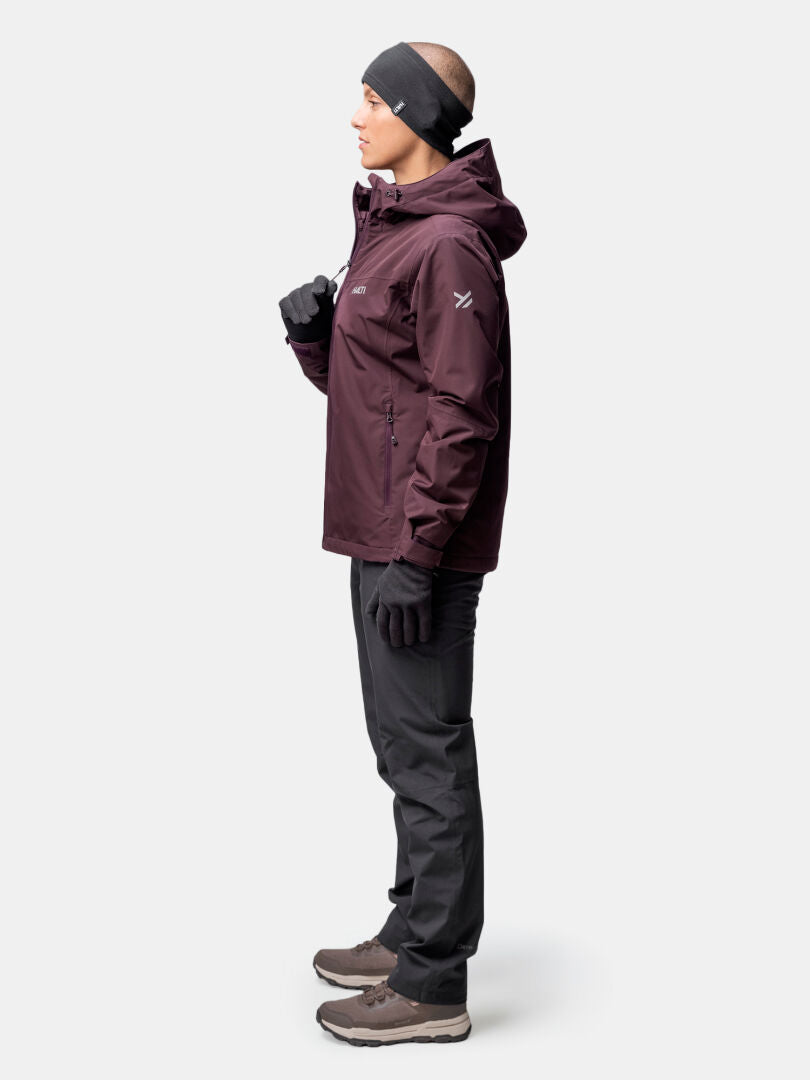 A woman in profile wears the Halti Forter DX Warm Shell Jacket in maroon, with black pants, gloves, boots, and a headband.