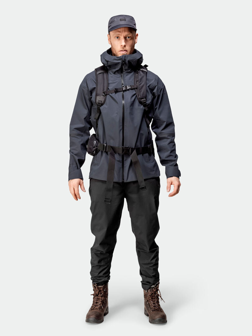 Person facing forward, wearing Halti Kajana 3L DX Shell Jacket, cargo pants, boots, cap, and a backpack with straps.