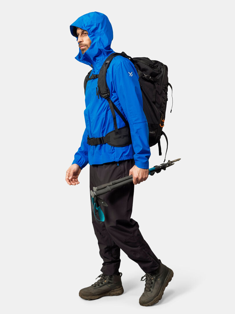A person in a Halti Pisarat 2,5L DX Shell Jacket, black pants, and boots carries a backpack and trekking poles.