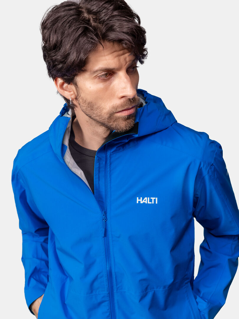 A man in a blue Halti Pisarat 2,5L DX Shell Jacket with hood looks left against a plain light background.