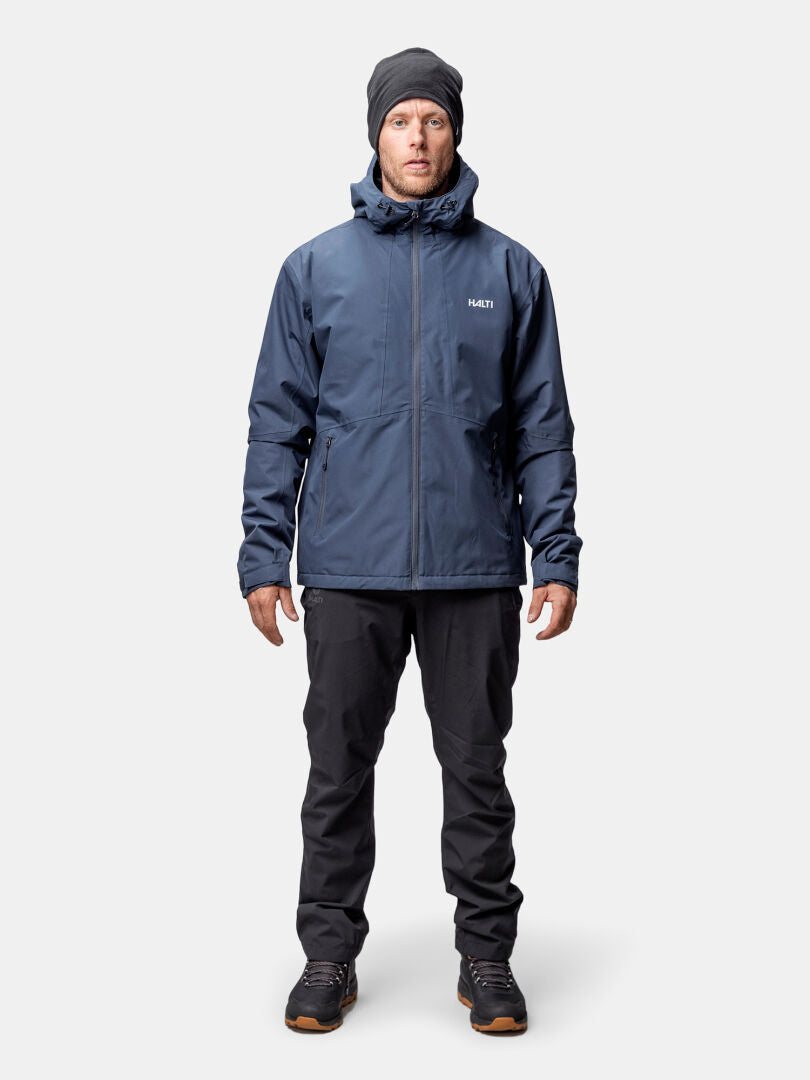 A man wears the Halti Forter DX Warm Shell Jacket in navy, black pants, beanie, and boots against a plain background.