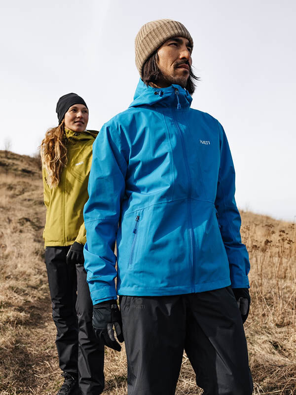 Two people in Halti Runko Beanies and outdoor jackets stand on a grassy hillside, looking ahead on a cloudy day.