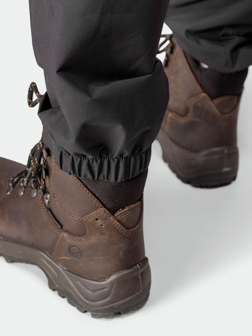 Close-up of a person wearing Halti Pisarat 2,5L DX Shell Pants Women's with elastic cuffs over dark brown hiking boots.