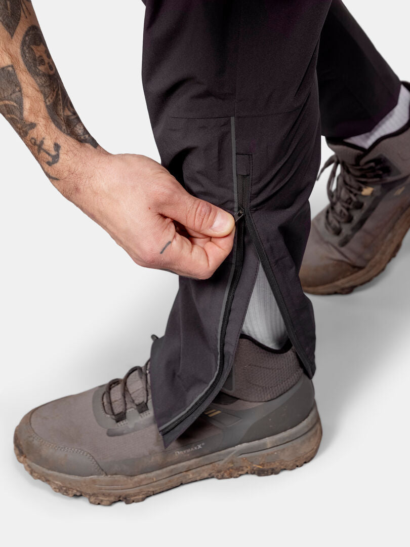 Someone unzips the lower leg of Halti Pisarat 2,5L DX Shell Pants Men's while wearing gray hiking shoes and white socks.