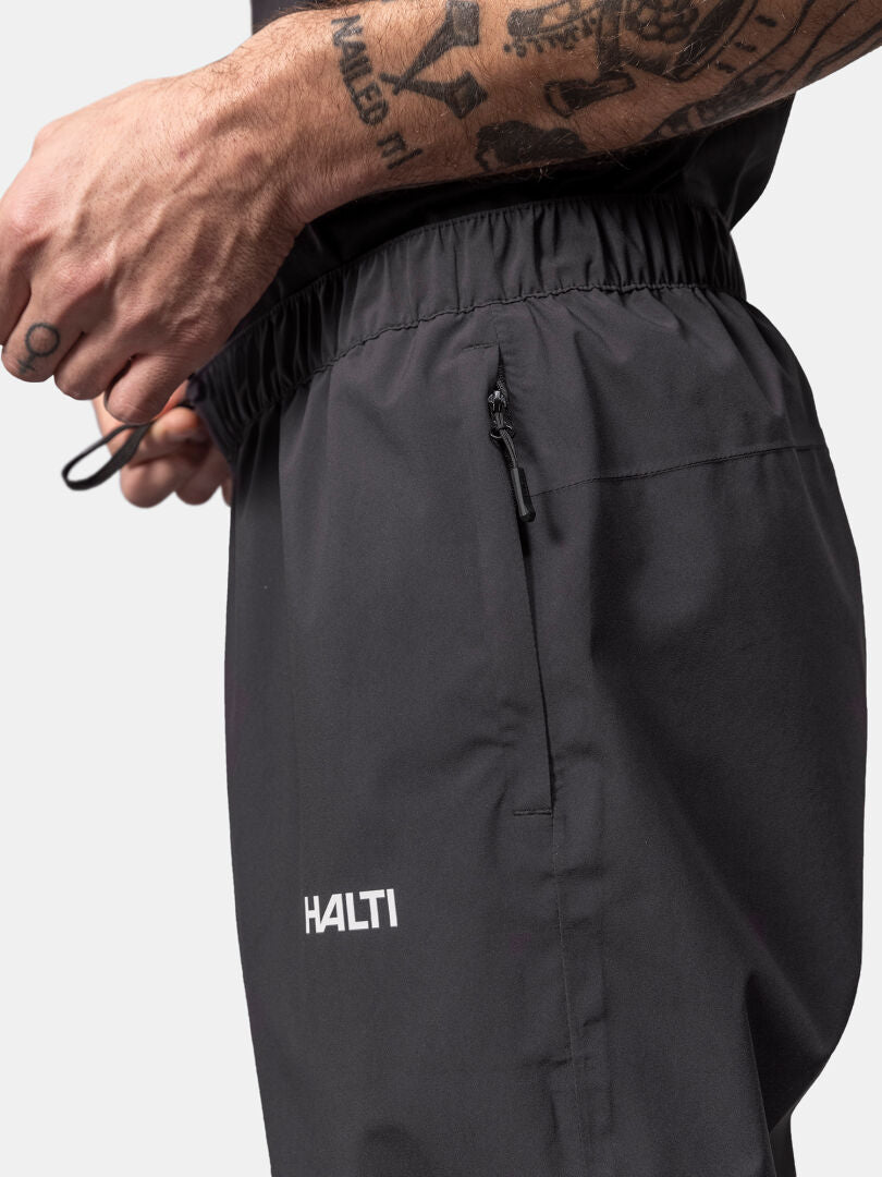 A tattooed person adjusts the drawstring on Halti Pisarat 2,5L DX Shell Pants with a zippered pocket.