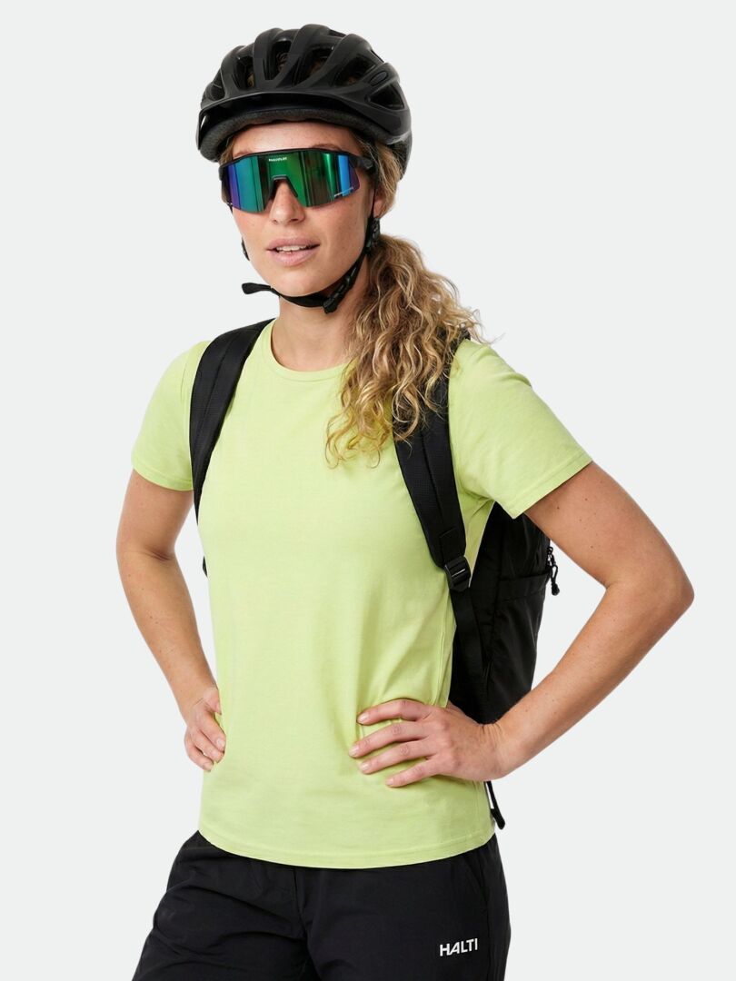 A woman in a Halti Kalteva T-shirt stands hands on hips outdoors, wearing a bike helmet and sunglasses.
