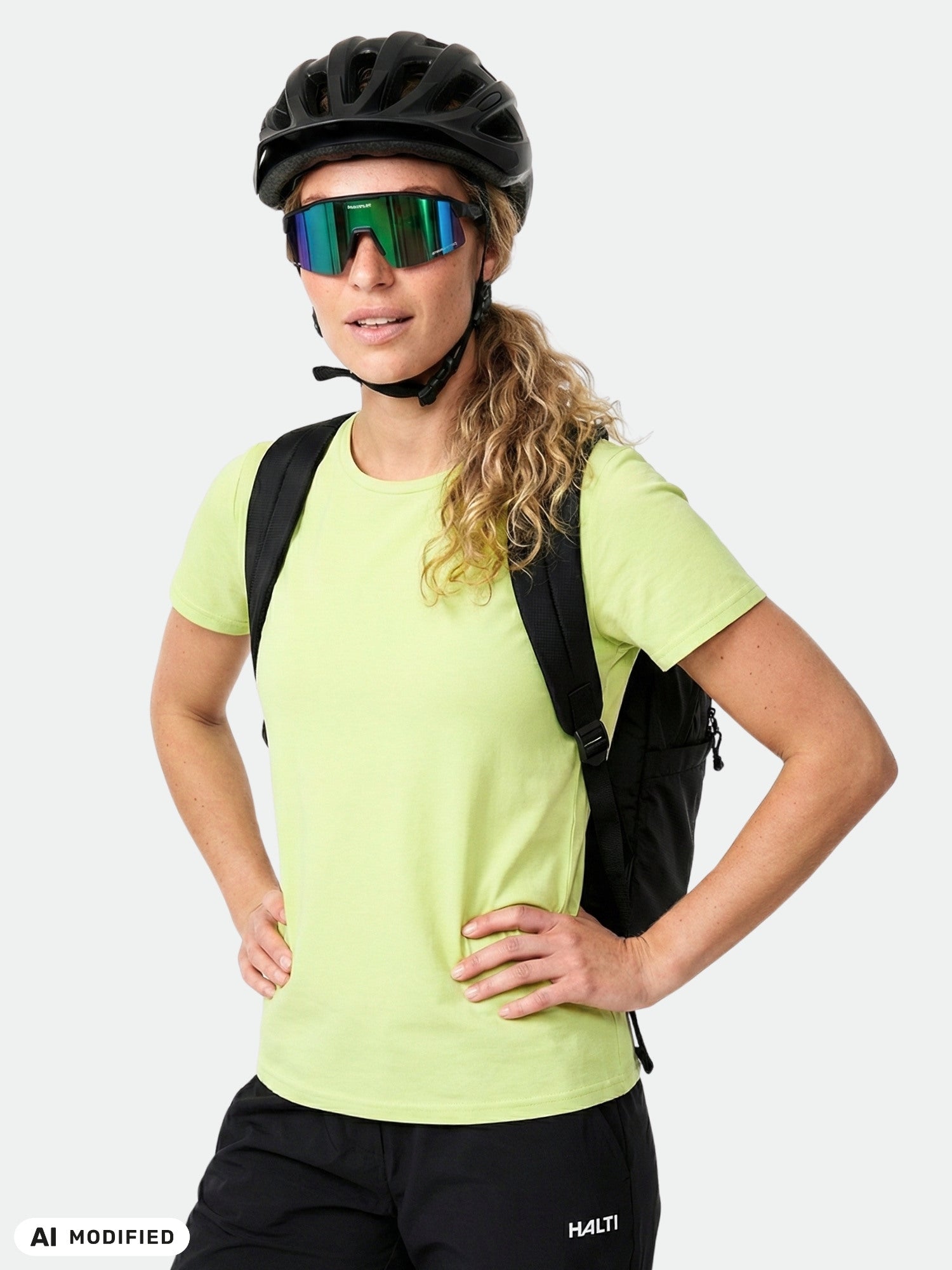 Woman wearing Halti Kalteva T-shirt, black helmet, reflective shades, lime green shirt, and backpack, hands on hips.