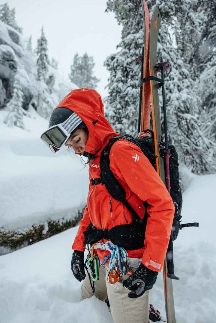 A woman in Halti Carvey II DX Ski Pants stands in snow with skis and gear, snowy trees behind her.