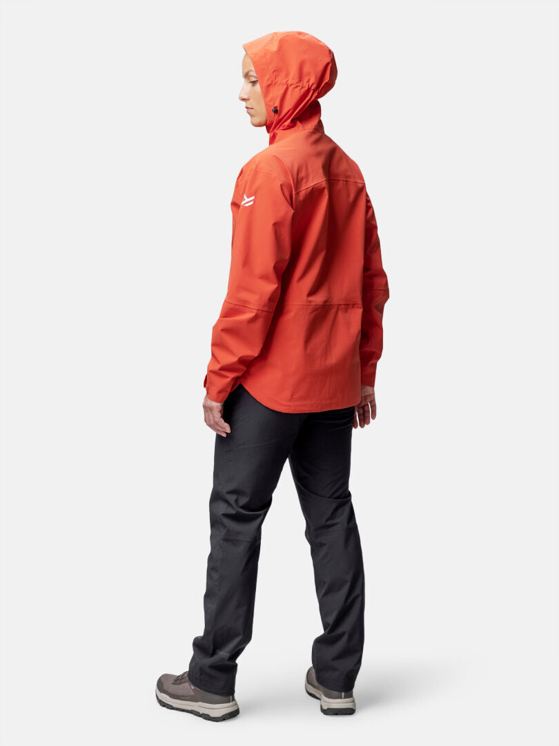 Woman wearing Halti Staala DrymaxX Shell Jacket in orange and dark pants stands facing left on a white background.
