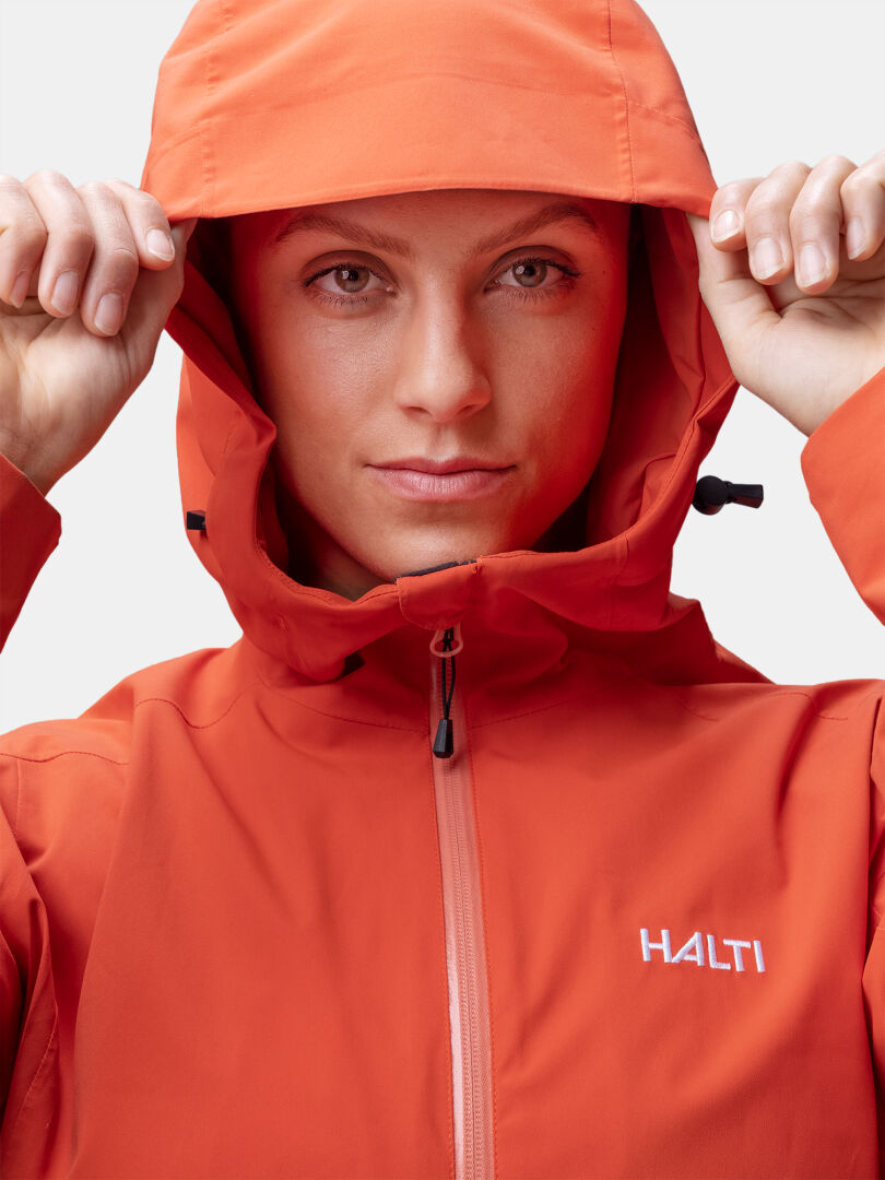 Model wearing Halti Staala DX Shell Jacket Women's in orange, hood up, facing forward, adjusting hood with both hands.