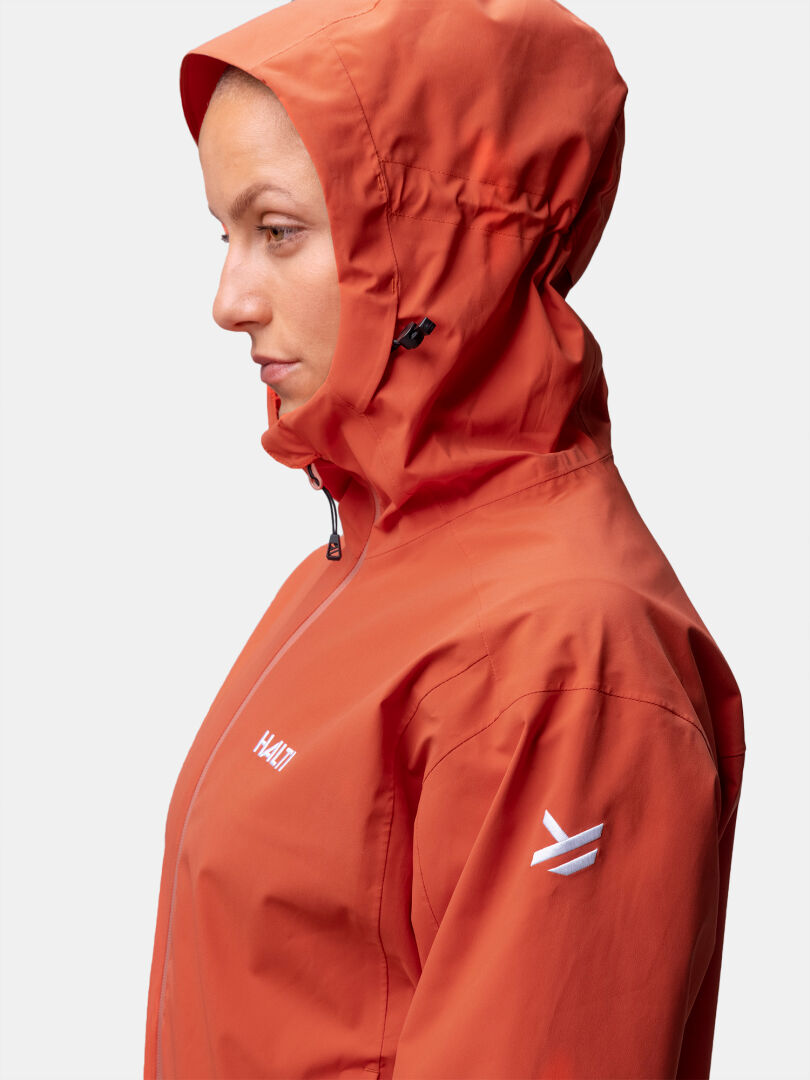 A woman in profile wears the Halti Staala DX Shell Jacket, an orange hooded outdoor style with a front zipper.