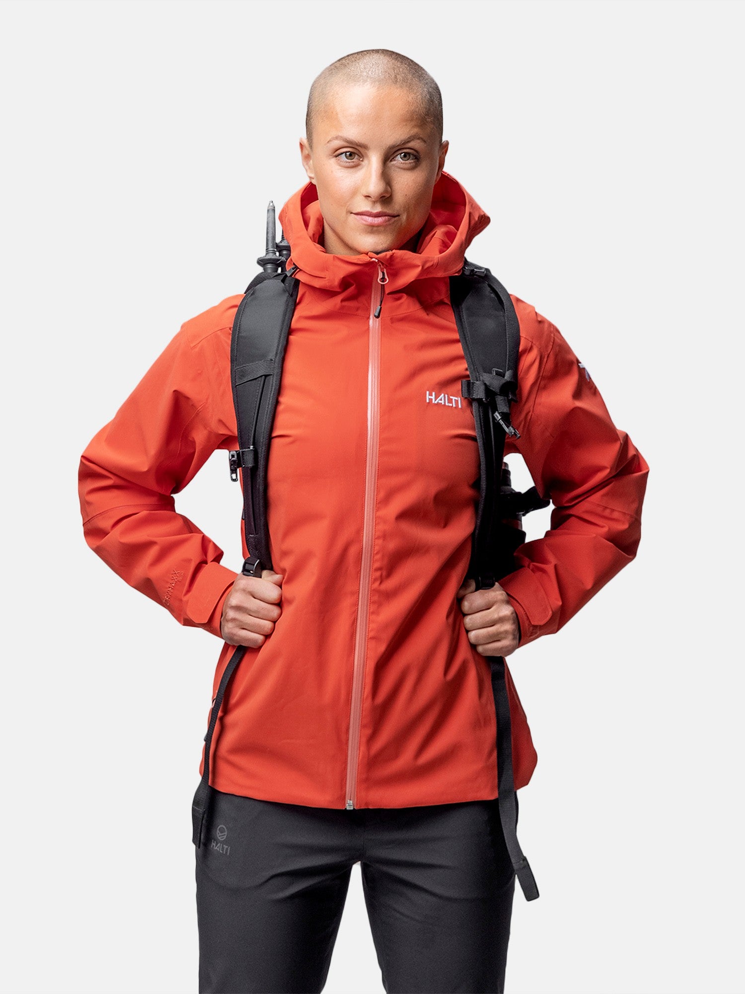 A person with a shaved head wears a red Halti Staala DrymaxX Shell Jacket and black backpack against a white background.