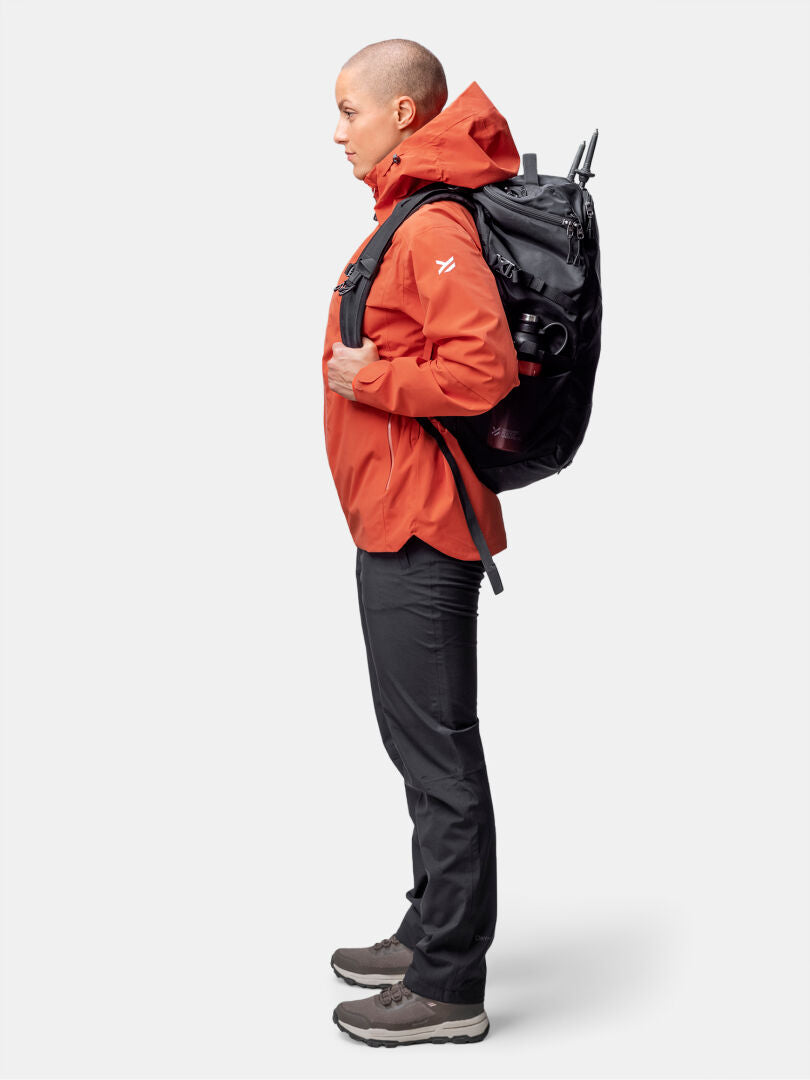 A woman in a Halti Staala DX Shell Jacket and black pants stands in profile with a large black backpack.