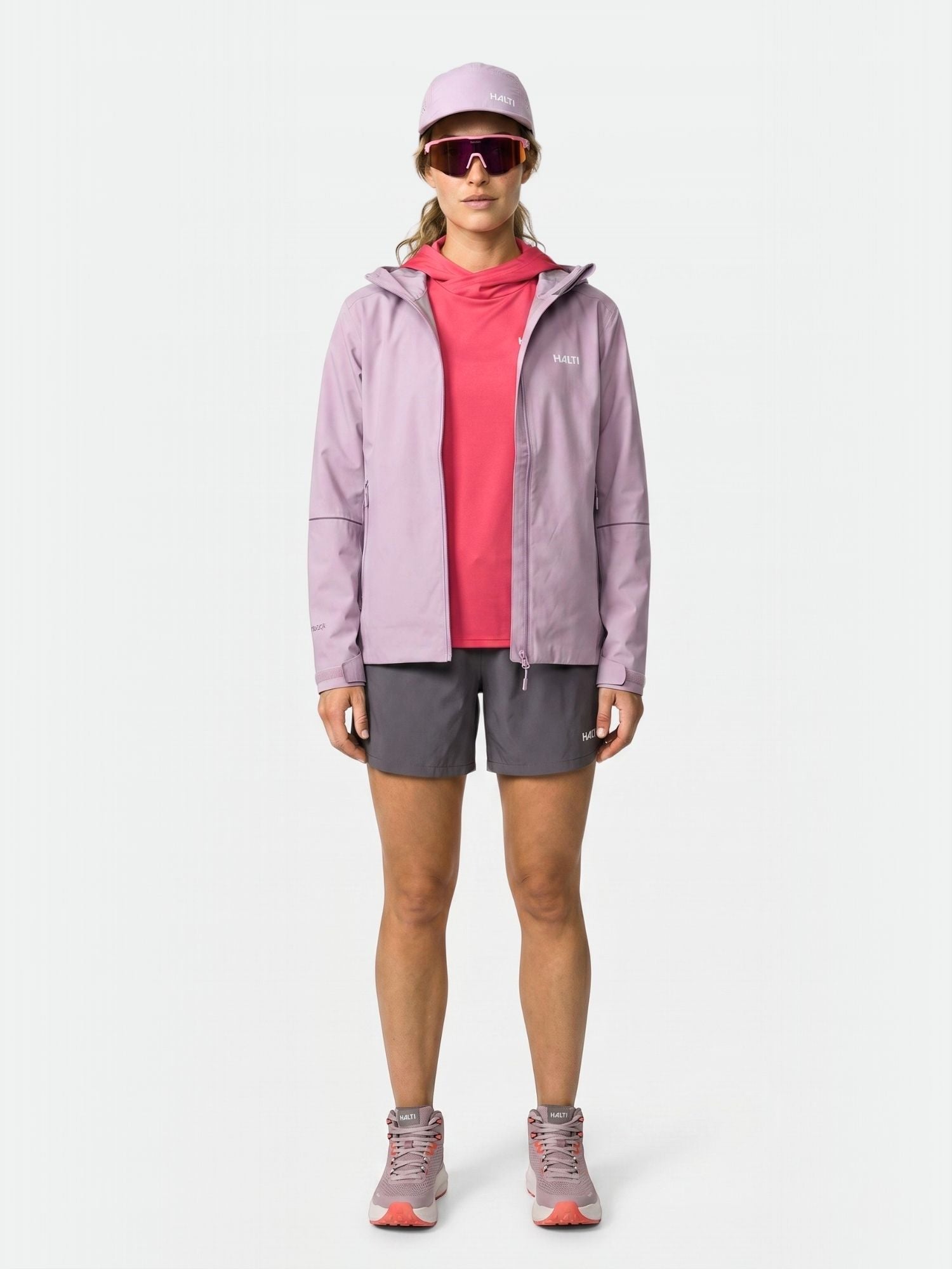 A woman faces forward wearing the Halti Staala DX Shell Jacket, pink shirt, gray shorts, cap, and sunglasses.