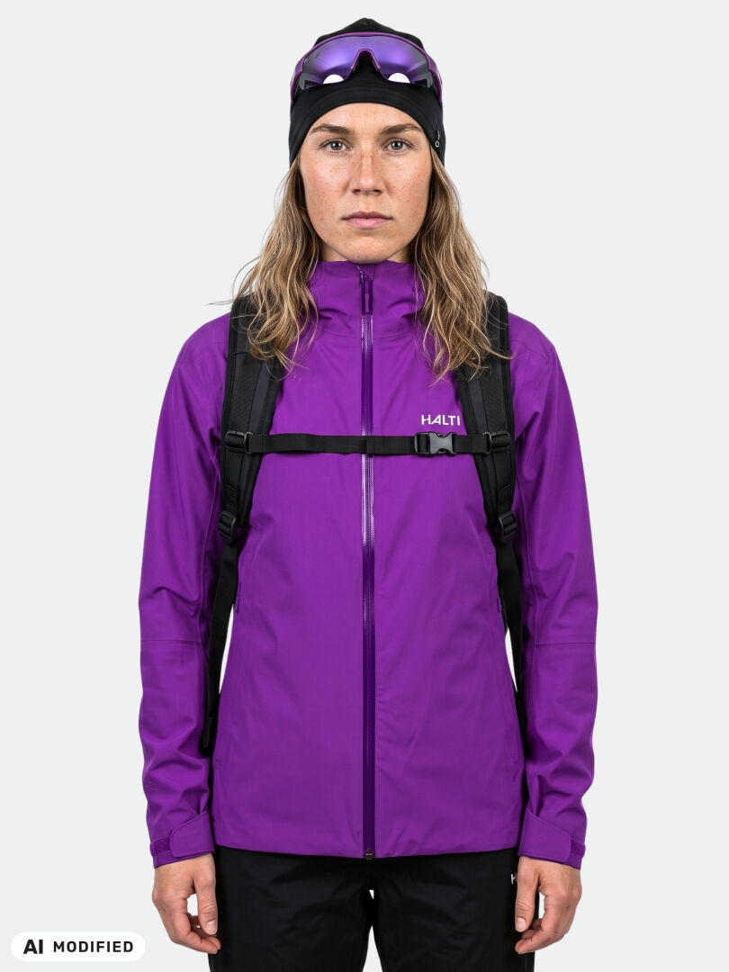 Woman in Halti Staala DX Shell Jacket, black backpack, and goggle hat stands against a plain light background.