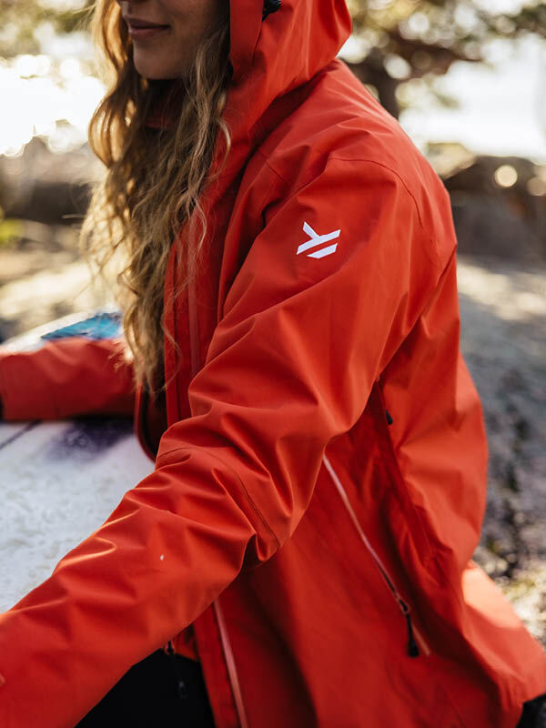 A person in a bright red Halti Staala DX Shell Jacket for women with a white logo sits outdoors near a table.