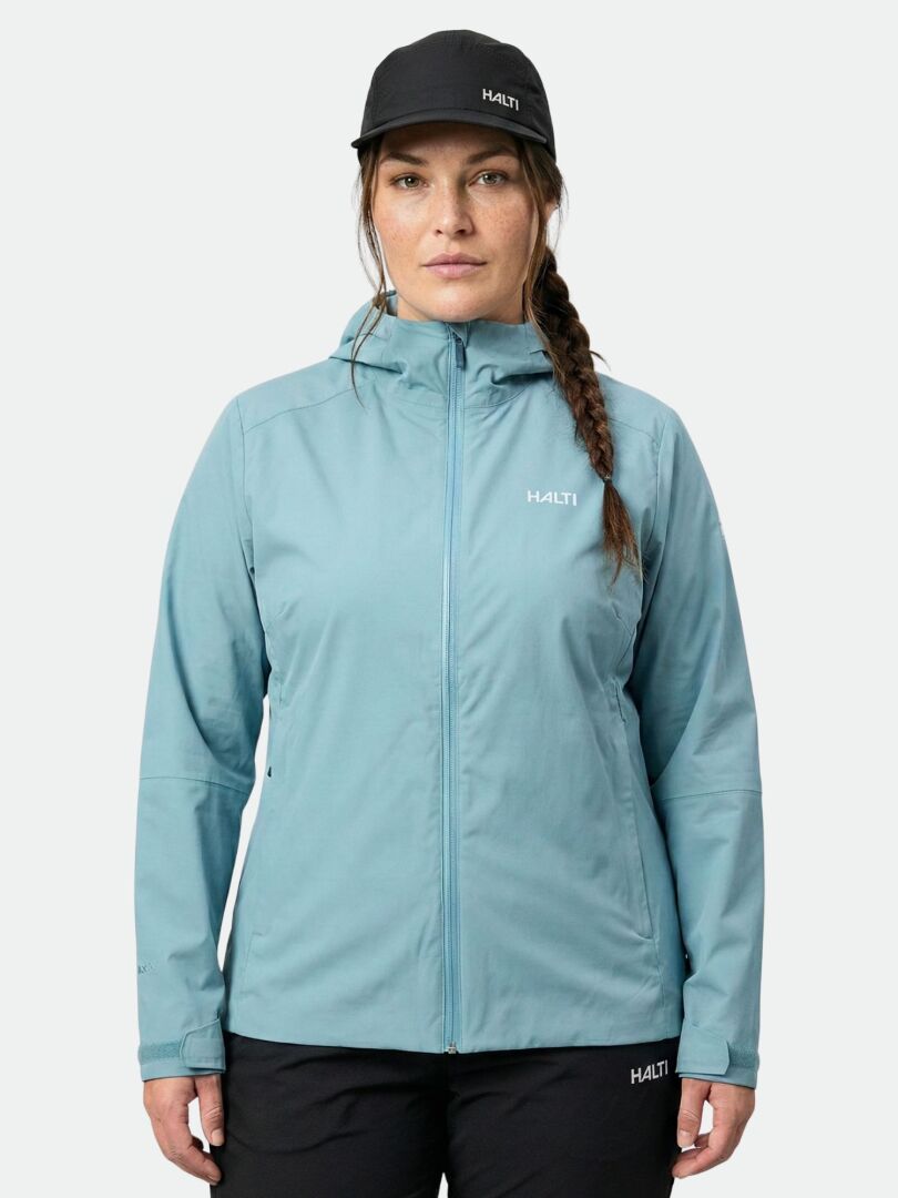 A woman wears a light blue Halti Staala DX Shell Jacket Women's Plus and a black Halti cap against a plain background.