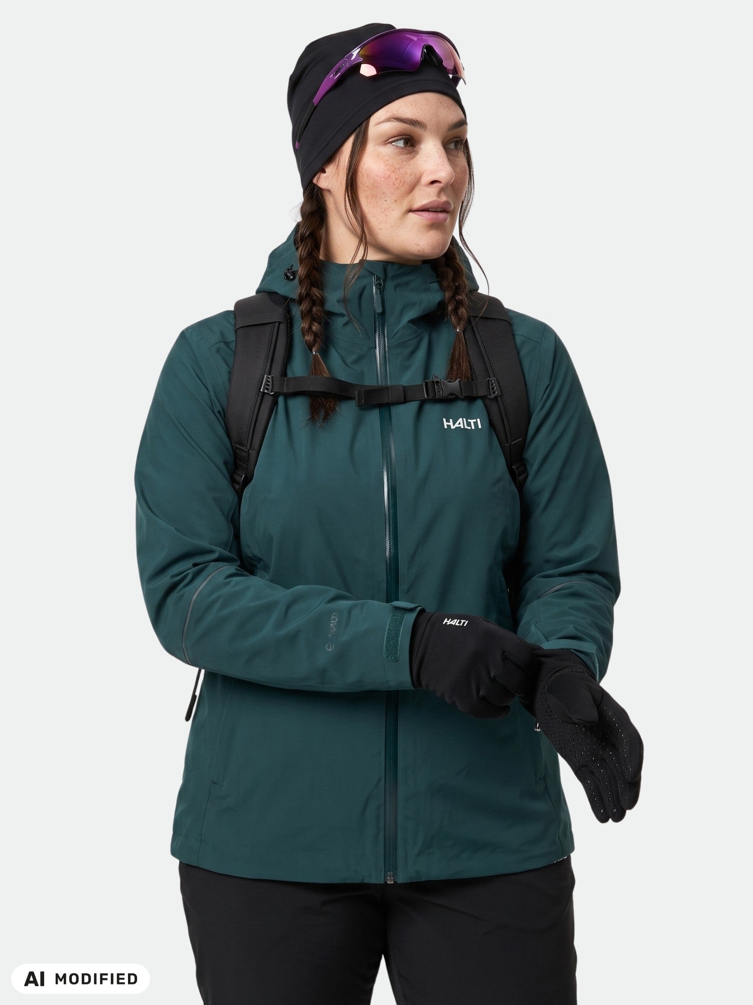 Person wearing Halti Staala DX Shell Jacket Women’s Plus, black gloves, beanie, sunglasses, and backpack looks aside.