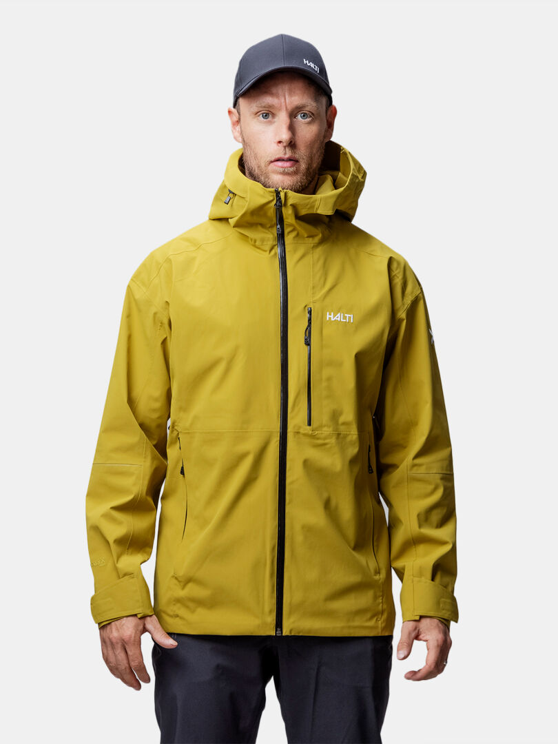 A man in a yellow Halti Staala DX Shell Jacket Men's and dark cap stands facing forward against a white background.
