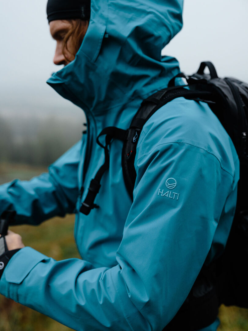 Man wearing Halti Staala DX Shell Jacket and backpack outdoors in fog, facing sideways.