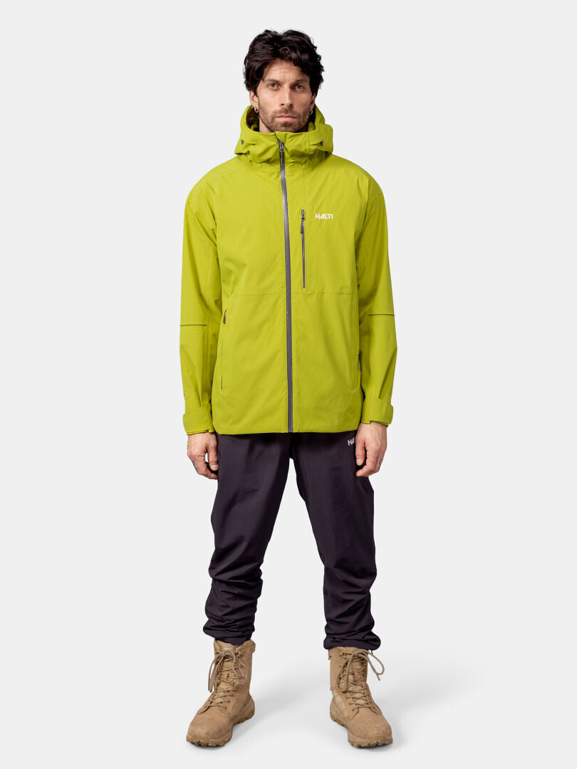A man wears the Halti Staala DX Shell Jacket in bright green, with dark pants and tan boots against a plain background.