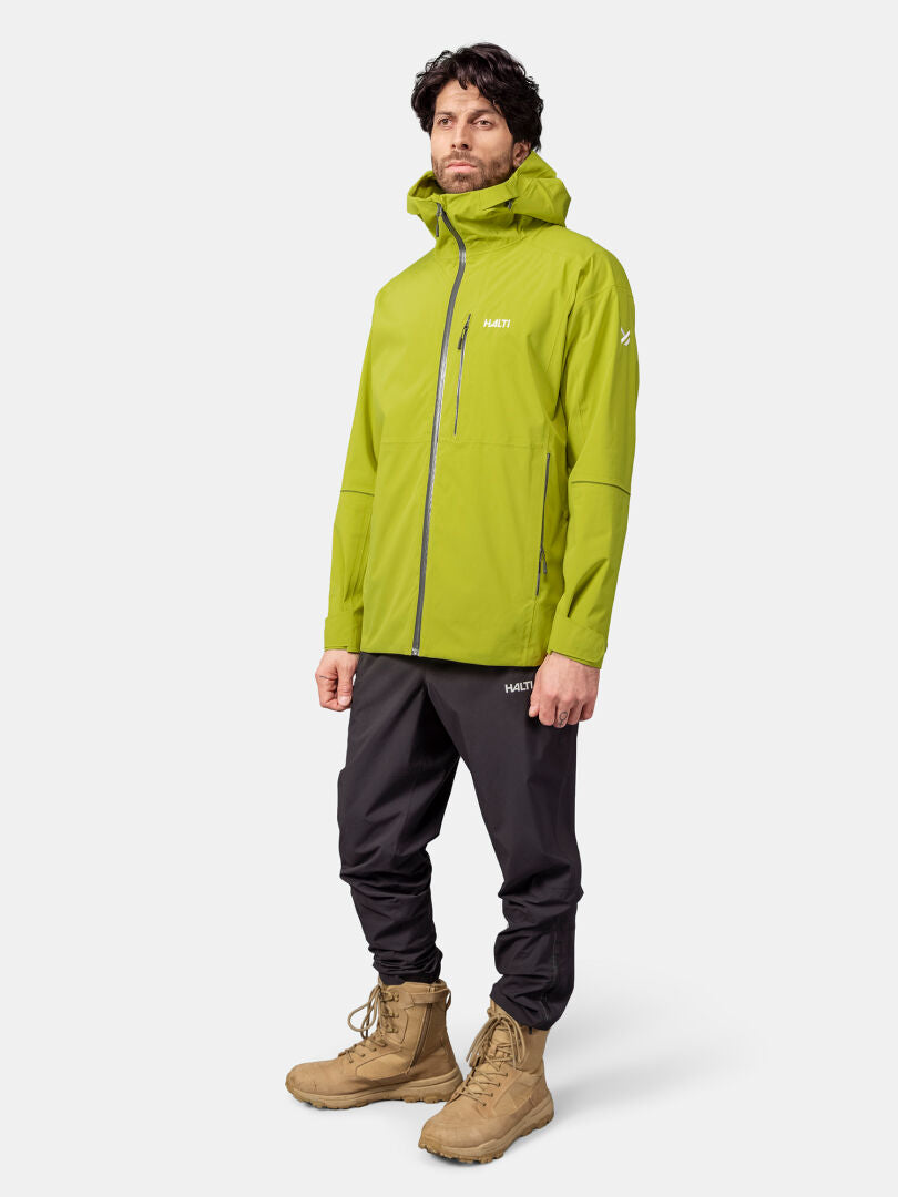 A man wears a Halti Staala DX Shell Jacket in lime green, black pants, and tan boots against a plain background.
