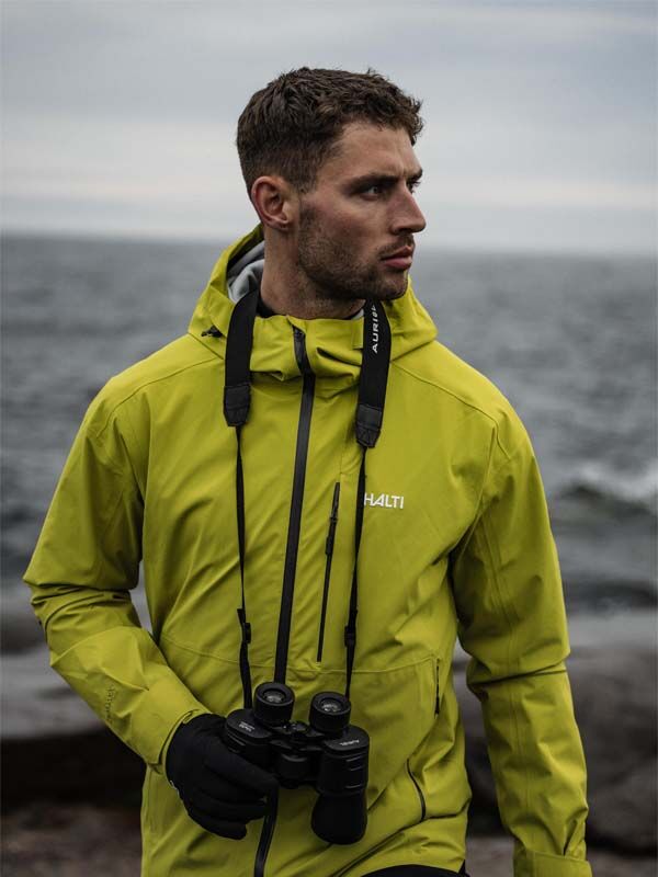 A man in a Halti Staala DX Shell Jacket stands with binoculars by a rocky shore on a cloudy day.