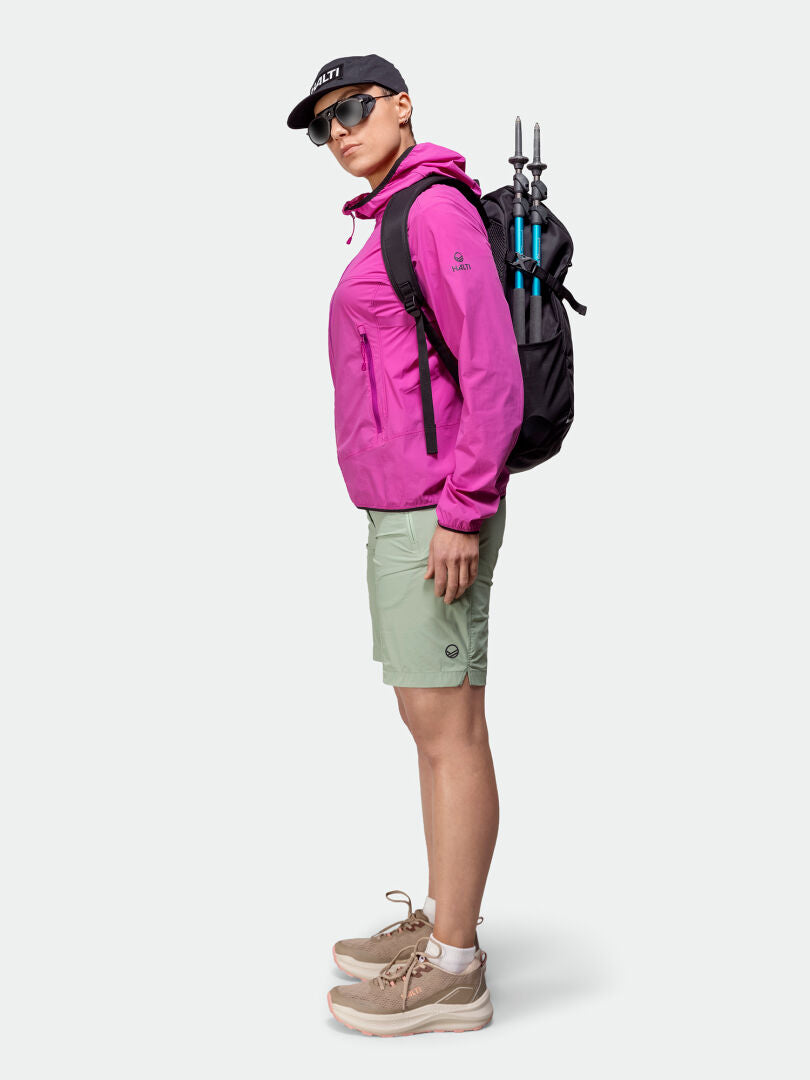 Woman in Halti Pallas Cool X-stretch Jacket (pink), green shorts, sneakers, sunglasses, cap, backpack, trekking poles.