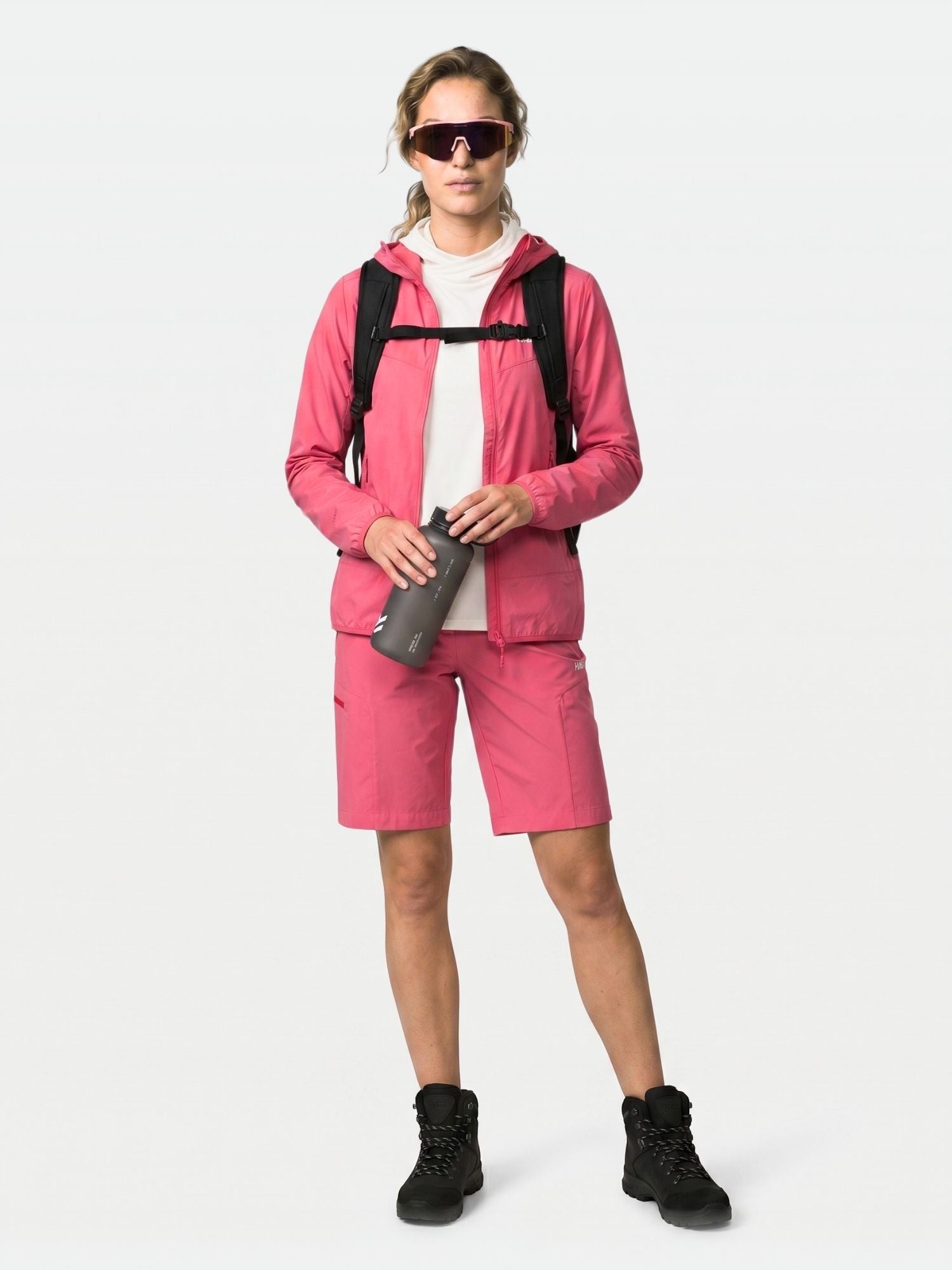 A woman in a Halti Pallas Cool X-stretch Jacket, shorts, sunglasses, black boots, backpack, and holding a water bottle.