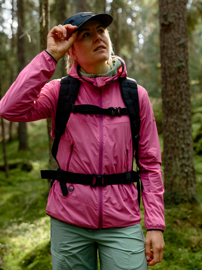 Woman wearing the Halti Pallas Cool X-stretch Jacket adjusts her cap with a backpack in a green forest.
