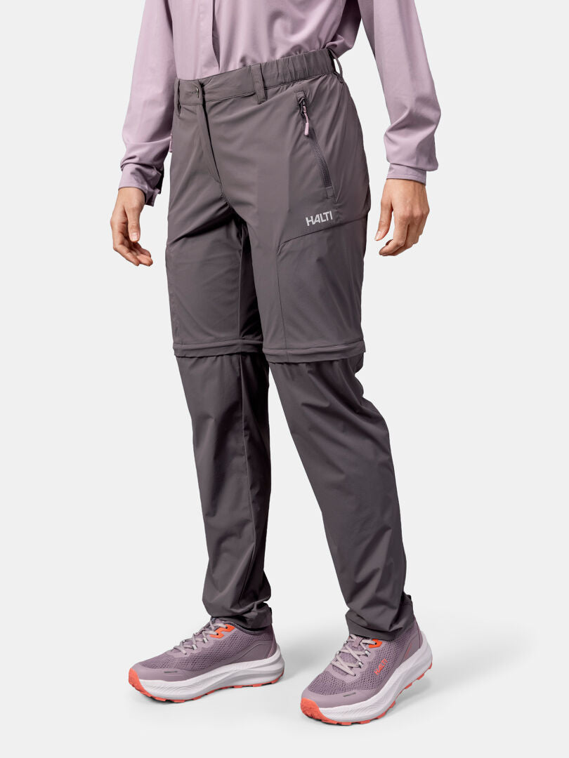 A woman in Halti Pallas Cool Stretch Zip-off Pants, a long-sleeve shirt, and sneakers stands against a plain background.