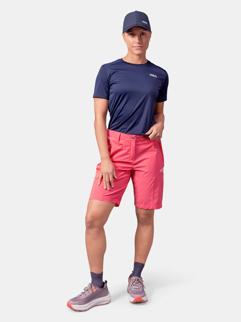 Person wearing a navy cap, navy t-shirt, Halti Pallas Cool Shorts Women's in pink, dark socks, and gray sneakers.