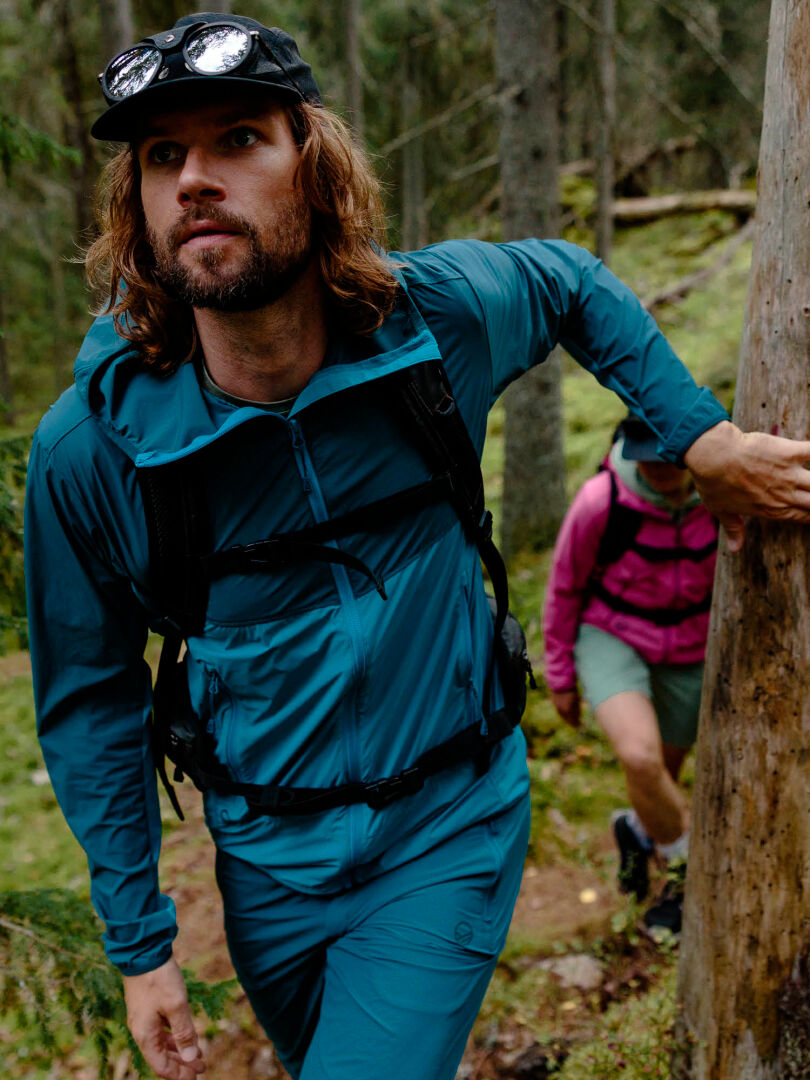 A man in a Halti Pallas Cool X-stretch teal jacket and black cap hikes uphill, followed by another in a pink jacket.