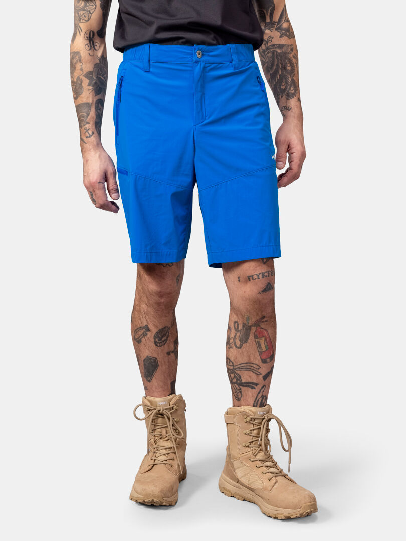 Person in Halti Pallas Cool X-stretch Shorts (blue), tan combat boots, black shirt, tattooed arms and legs.