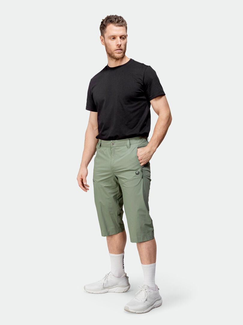 A man stands wearing Halti Pallas Cool X-stretch Capri Pants, a black t-shirt, white socks, and white sneakers.