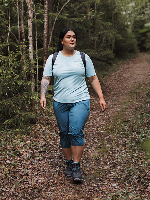 A woman wearing Halti Pallas Cool X-stretch Capri Pants walks with a backpack on a forest trail among trees.
