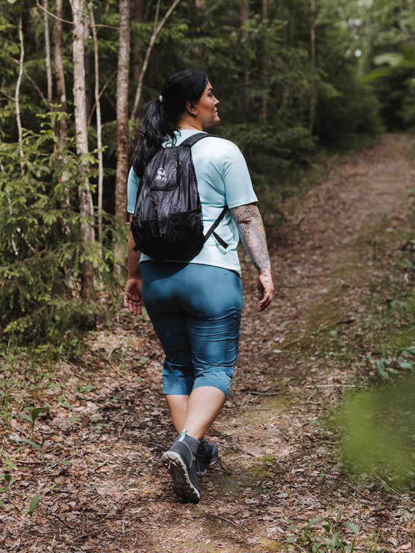 A person in Halti Pallas Cool X-stretch Capri Pants walks a dirt trail through a lush, green forest.