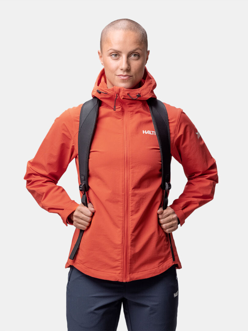 A woman wears the Halti Finholma X-stretch Jacket in orange with a black backpack, standing against a white background.