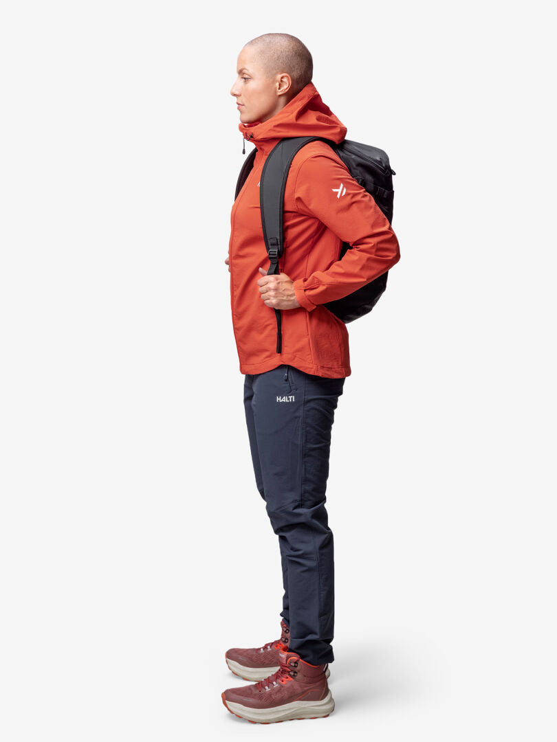 A person in profile wears the Halti Finholma X-stretch Jacket Womens in red, with a black backpack, dark pants, and red boots.