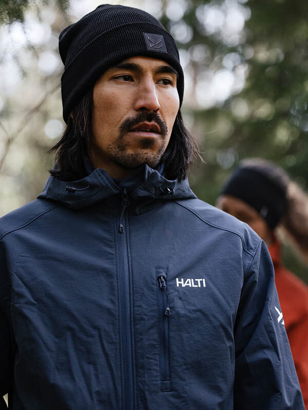 A man in a dark jacket wears a Halti Vataja Beanie outdoors, with another person blurred in the background.