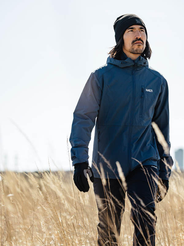 Man in a Halti Finholma X-stretch Jacket, black beanie, and gloves walks through tall dry grass on a sunny day.