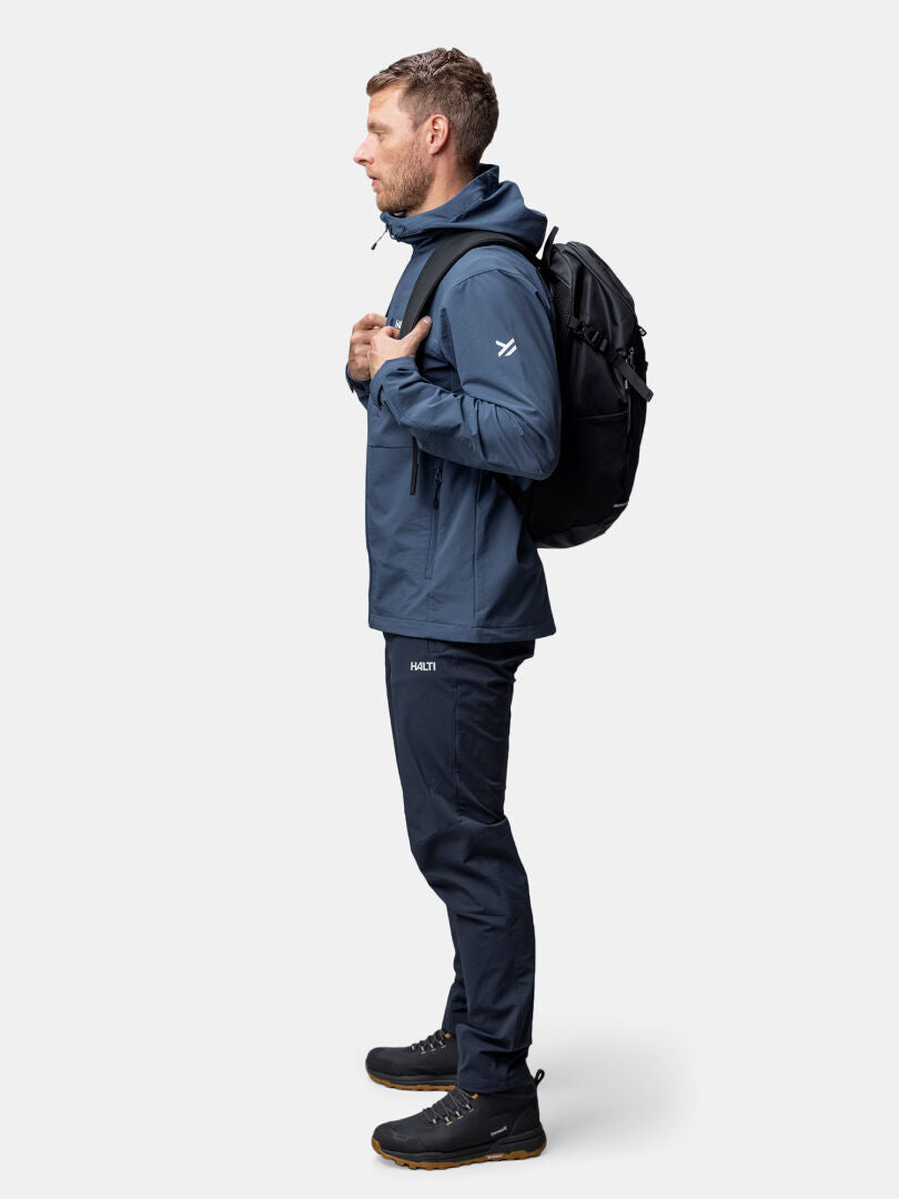 A man in the Halti Finholma X-stretch Jacket stands in profile, with a black backpack and shoes against a plain background.