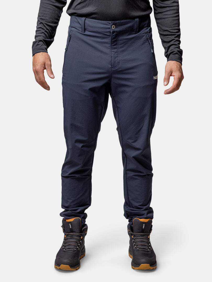 A person in Halti Finholma X-stretch Pants, black long-sleeve top, and hiking boots with brown soles stands upright.
