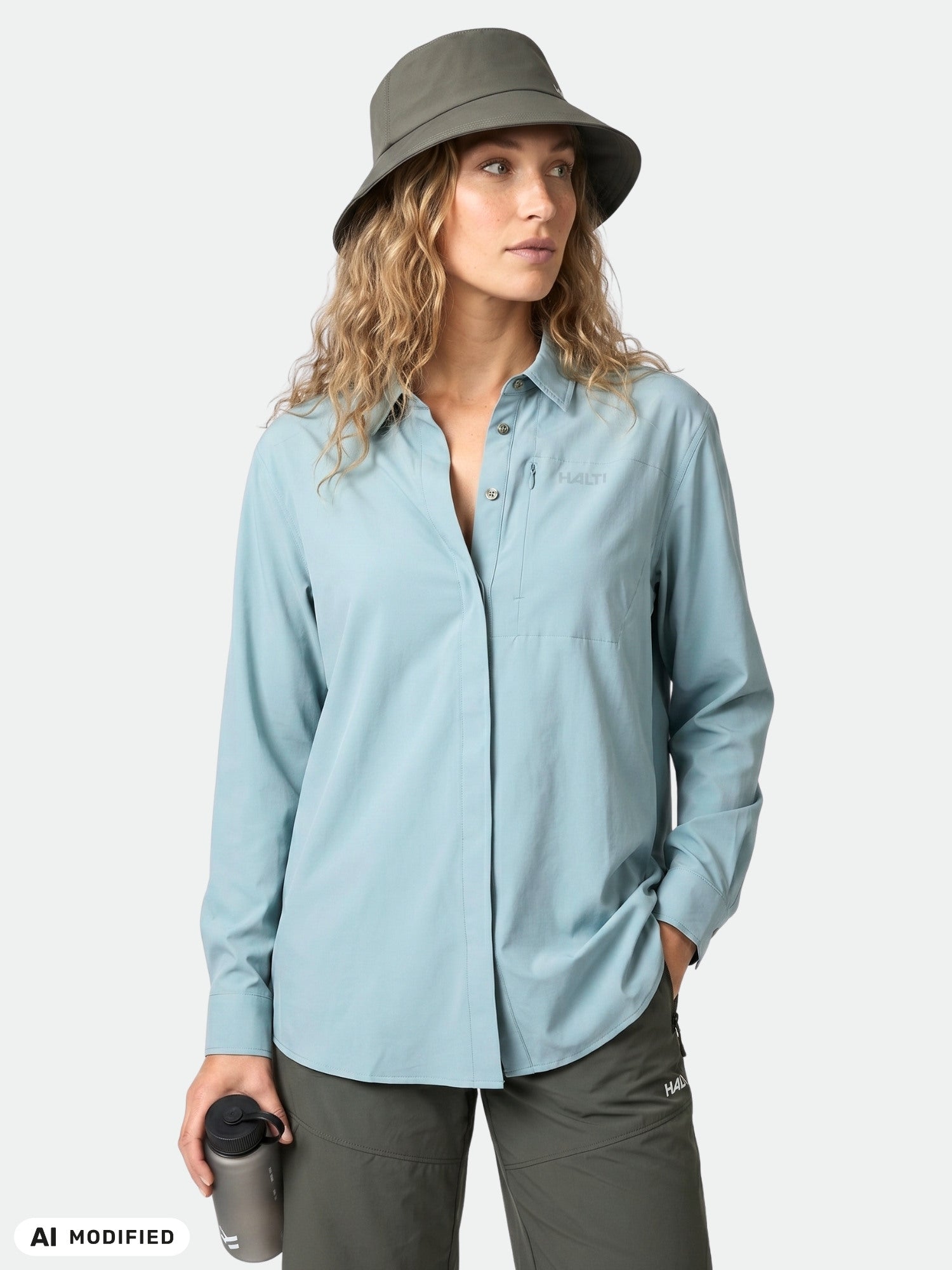 Woman in Halti Samoy Longsleeve Trekking Shirt, olive hat, green pants holds travel mug and looks aside.