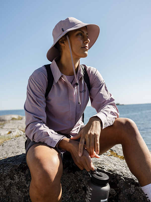 A woman in a Halti Samoy Longsleeve Trekking Shirt with UPF 90+ sits by the water, gazing into the distance.