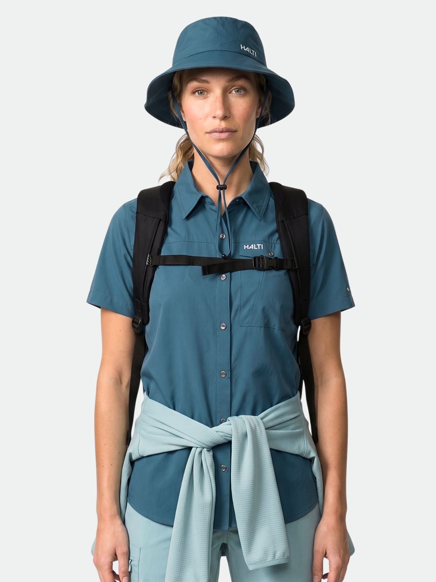 A woman in a Halti Samoy Trekking Shirt, bucket hat, backpack, and jacket tied at the waist, facing forward.